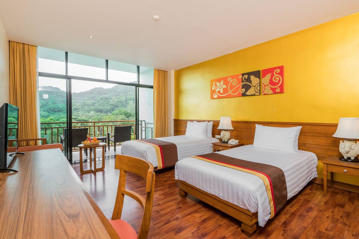 Ein Hotelzimmer mit zwei Betten, einem Holztisch, einem Stuhl und einem Balkon mit Blick auf Grünflächen.