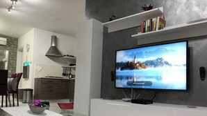 Un salon moderne avec un téléviseur à écran plat fixé au mur, une table basse en verre et un coin cuisine à l’arrière-plan.
