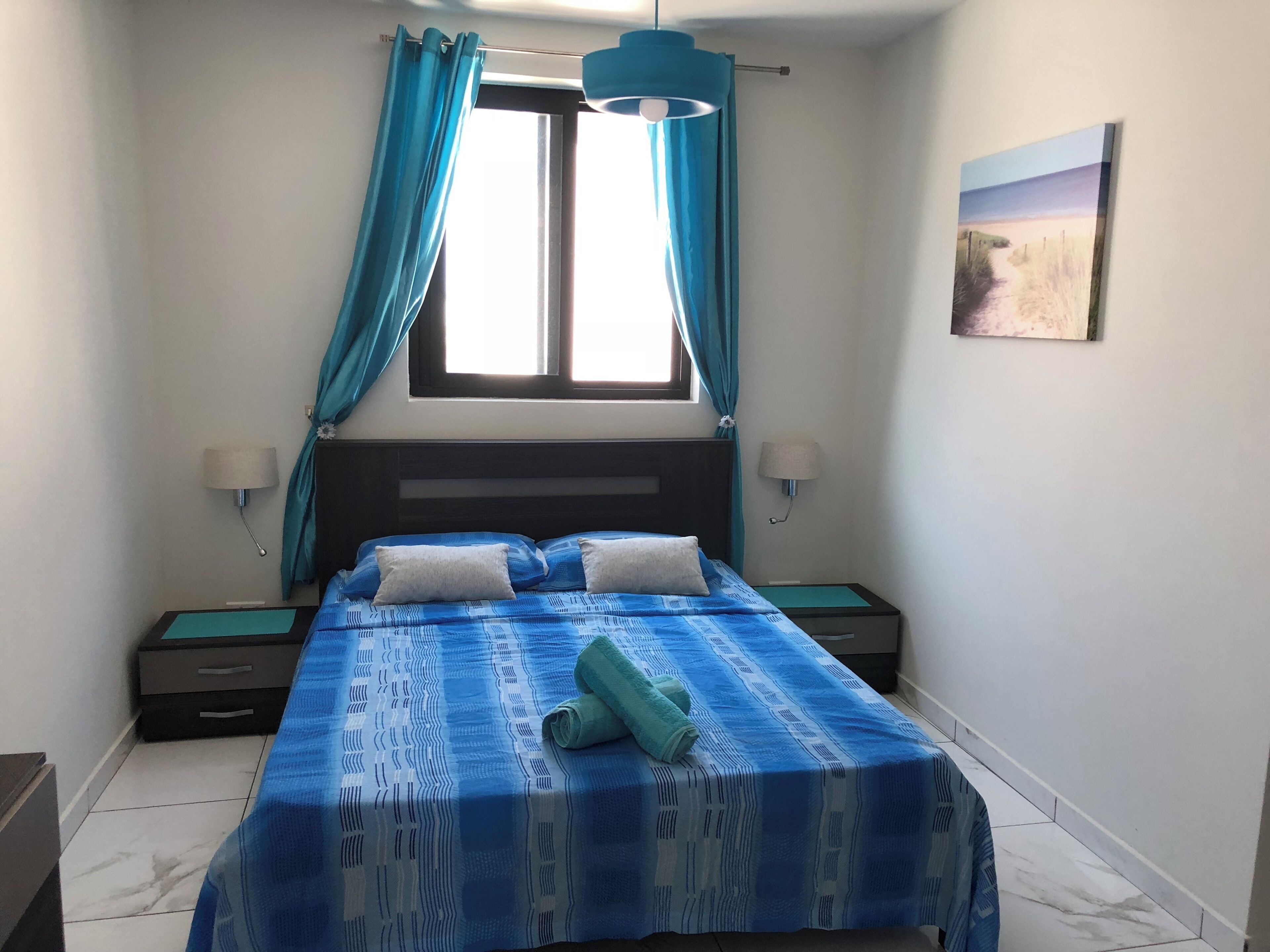 Une chambre à coucher comprenant un lit, deux tables de chevet, une fenêtre avec des rideaux, une lampe fixée au mur et un tableau.