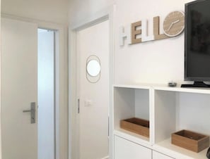 Ett rum med en vit dörr, en väggklocka, en skylt med texten ”HEJ” och en tv som står på ett vitt TV-bord.