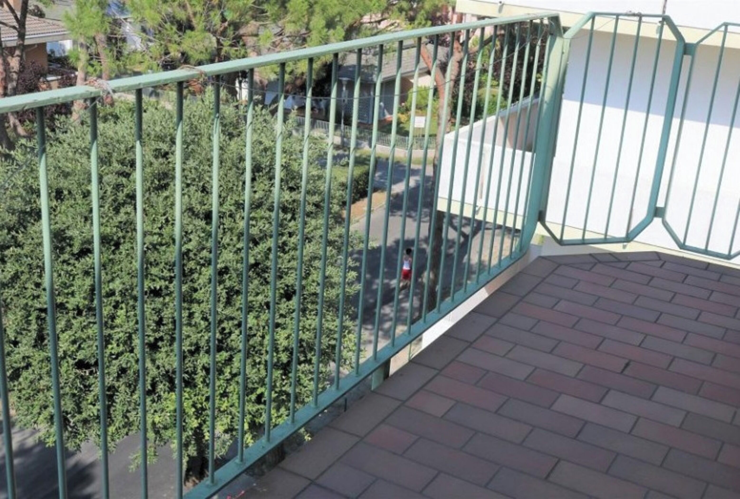 A balcony with a metal railing, brick flooring, and a view of a residential area with trees and houses.