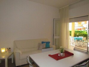 Ein kleiner, gut beleuchteter Raum mit einem weißen Sofa, einem kleinen Tisch und einem Fenster mit Blick auf einen Balkon und Gartenmöbeln.