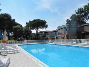 A swimming pool with lounge chairs and umbrellas, surrounded by a white fence and trees. - Homely Apartment With Private Garden (San Michele al Tagliamento)