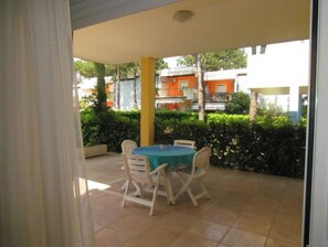 Eine Terrasse im Freien mit Tisch und Stühlen, umgeben von Grünpflanzen und einem Gebäude im Hintergrund.