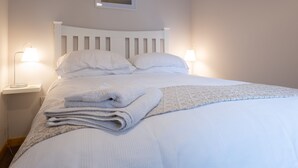 Una cama bien hecha con sábanas blancas, una toalla doblada y una manta. Hay dos lámparas de noche a cada lado de la cama.