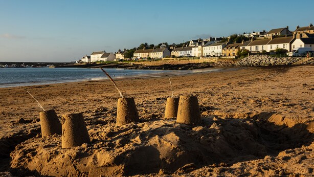 Una playa arenosa con cuatro castillos de arena y una hilera de casas al fondo.