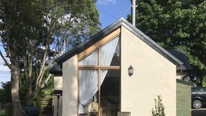 Una casa pequeña con un ventanal amplio, un banco de madera y un jardín con plantas y una jardinera de madera.