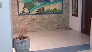Ein Korridor mit Fliesenboden, einem großen Wandgemälde, das eine Landschaft mit Bäumen und einem Haus zeigt, sowie einem Blumentopf mit getrockneten Blumen.