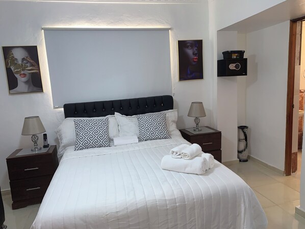 Un lit bien fait, avec du linge de lit blanc et des coussins à motifs, une table de chevet avec une lampe, une télécommande et un petit tiroir.