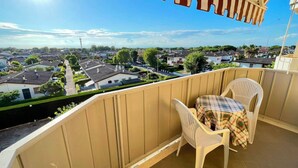 A balcony with a view of a residential area, a table with a plaid tablecloth, and two white plastic chairs. - Flat With Excellent Location (Caorle)