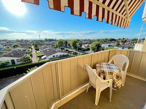 A balcony with a view of a residential area, a table with a plaid tablecloth, and two white plastic chairs.