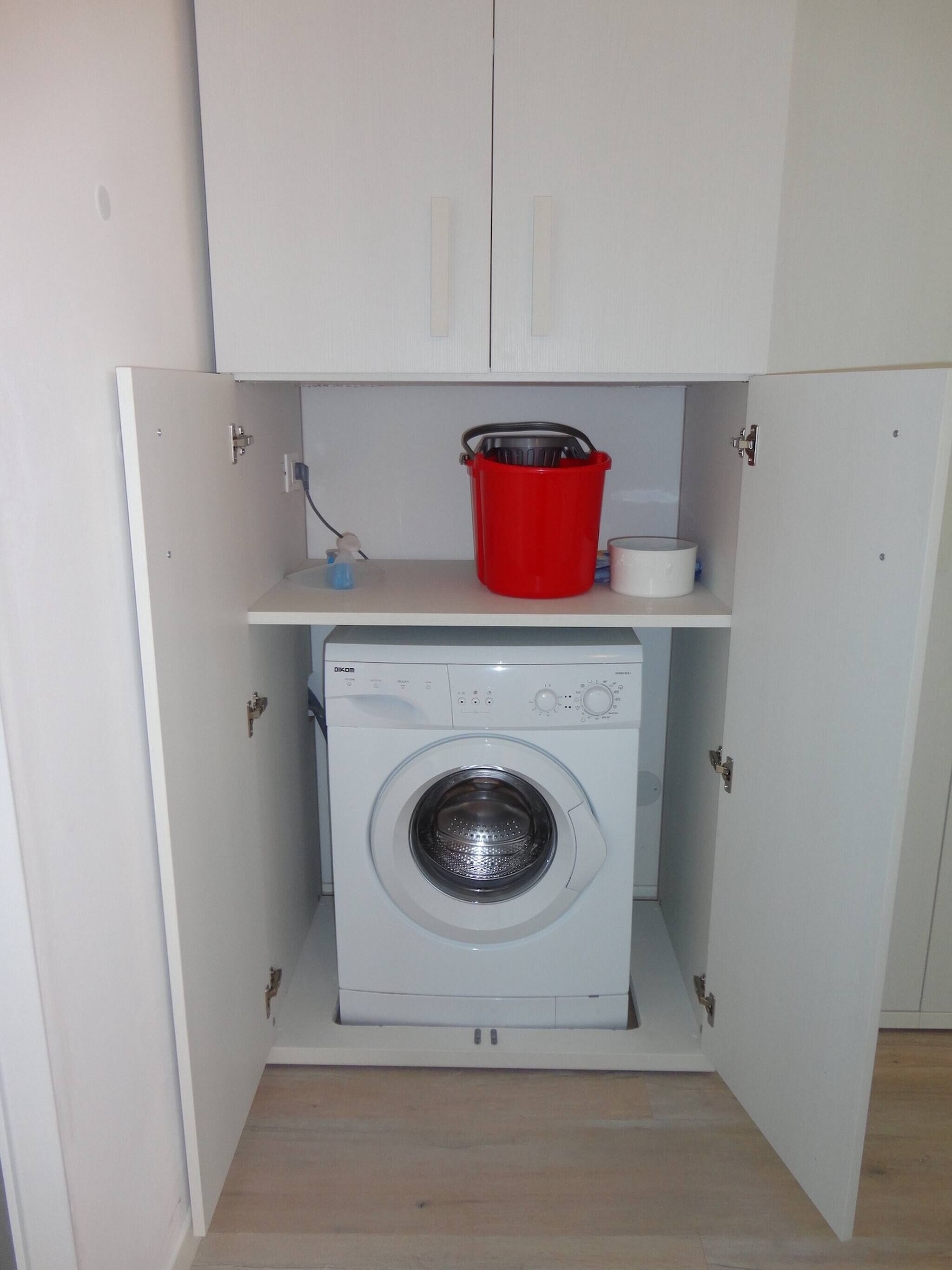 A compact laundry room with a front-loading washing machine, a red bucket, and a small shelf above the machine.