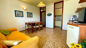 Un salon avec un canapé, une table à manger avec des chaises, un coin cuisine avec un évier et une cuisinière, et un luminaire fixé au plafond.