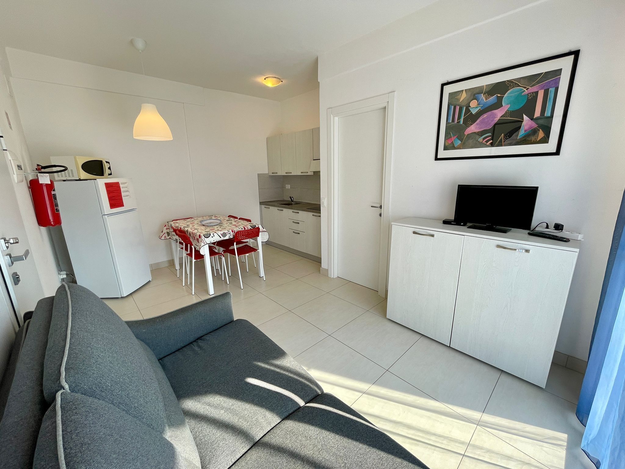 A compact living space with a sofa, a dining table with chairs, a kitchenette, a television, and a painting on the wall.
