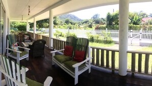 A covered porch with wooden chairs and a bench, white railings, and a view of a lush garden and mountains. - Casa Livingston - Luxury Villa - La Digue Seychelles (La Digue)
