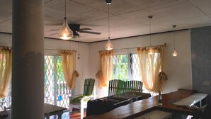 A dining area with a long wooden table, hanging pendant lights, and a view through a window with curtains. - Casa Livingston - Luxury Villa - La Digue Seychelles (La Digue)