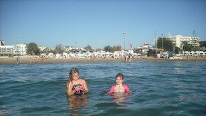 Two children in a body of water with a beach and buildings in the background.
