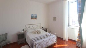Ein Schlafzimmer mit einem Metallbettgestell, einem Nachttisch, einem Sessel und einem Fenster mit Vorhängen.
