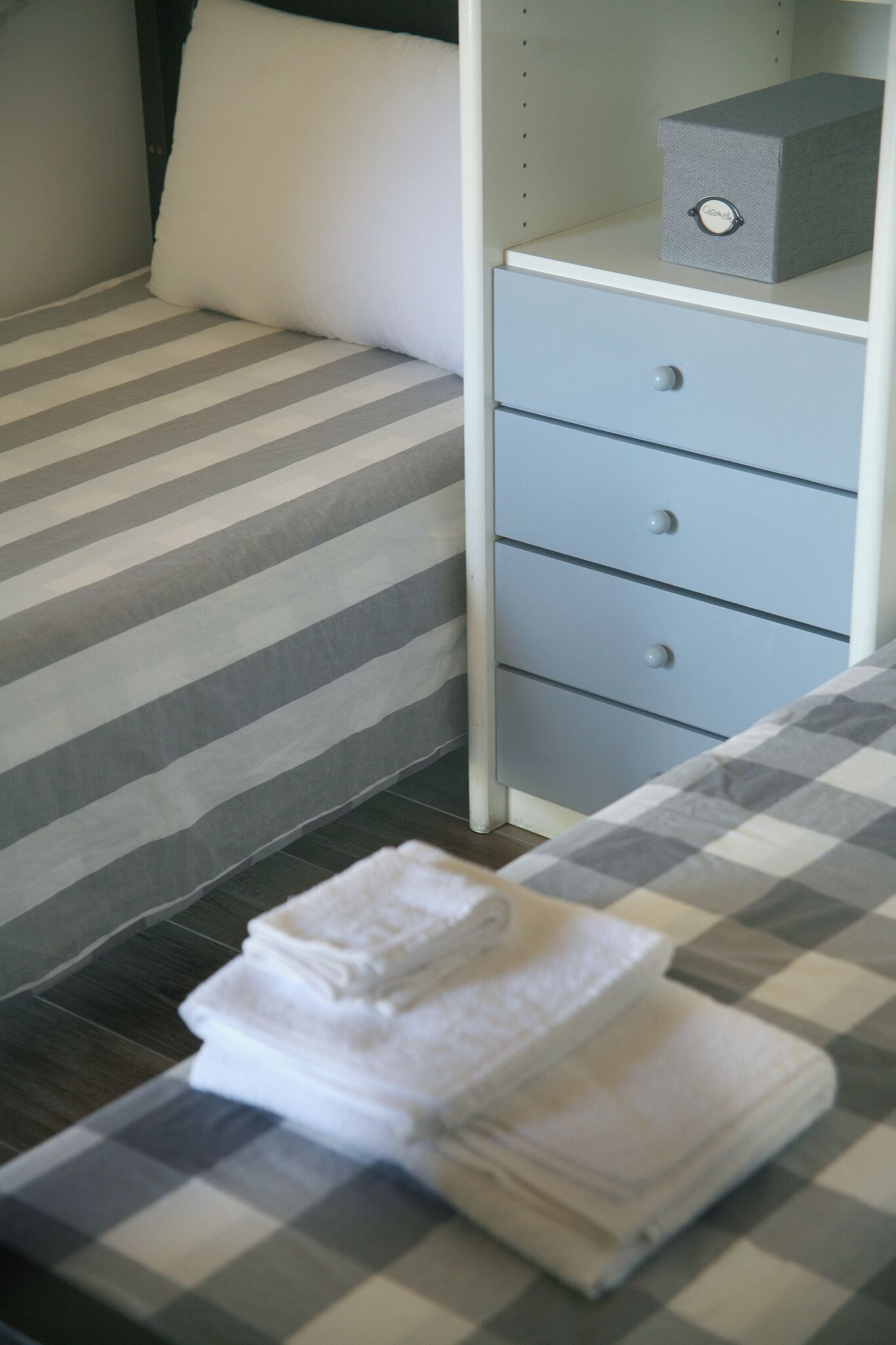 A neatly made bed with striped bedding, a drawer unit with three drawers, and a box on top.
