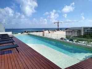A rooftop infinity pool with wooden deck, clear blue water, and a view of the ocean and construction cranes in the distance. - Luxurious Charming Studio-terrace With Pool-gym (Playa del Carmen)