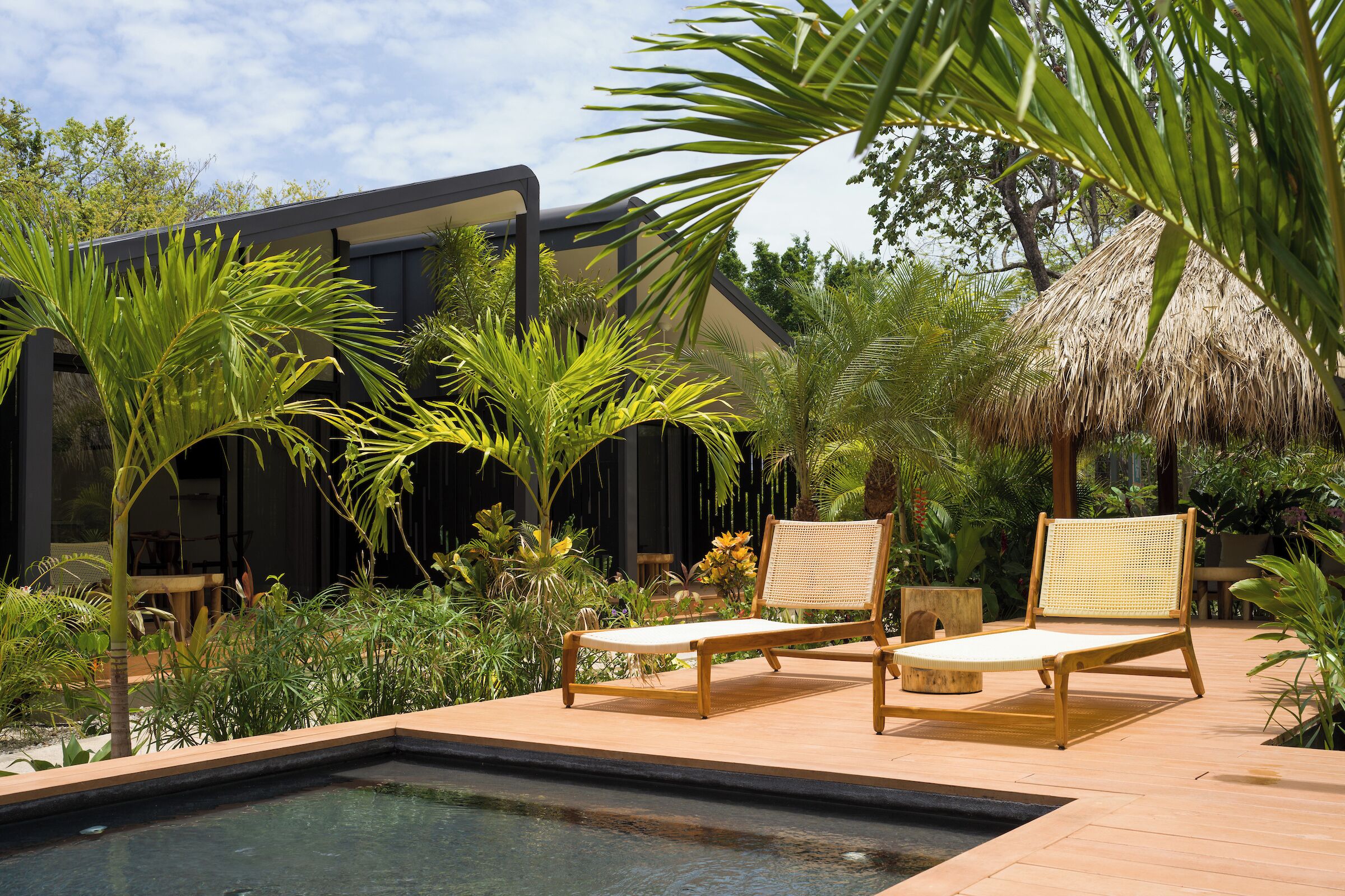 A poolside area with two wooden lounge chairs, a thatched-roof structure, and lush tropical plants.