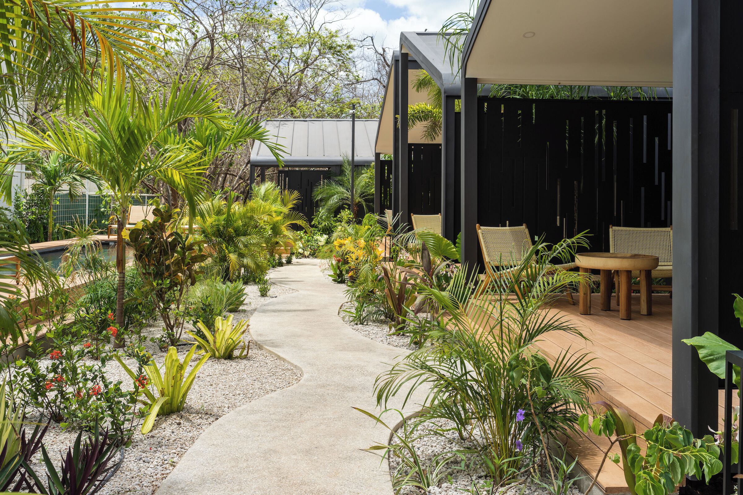 Un sentier bordé de différentes plantes qui mène à un bâtiment moderne doté d’un espace extérieur aménagé pour s’asseoir.