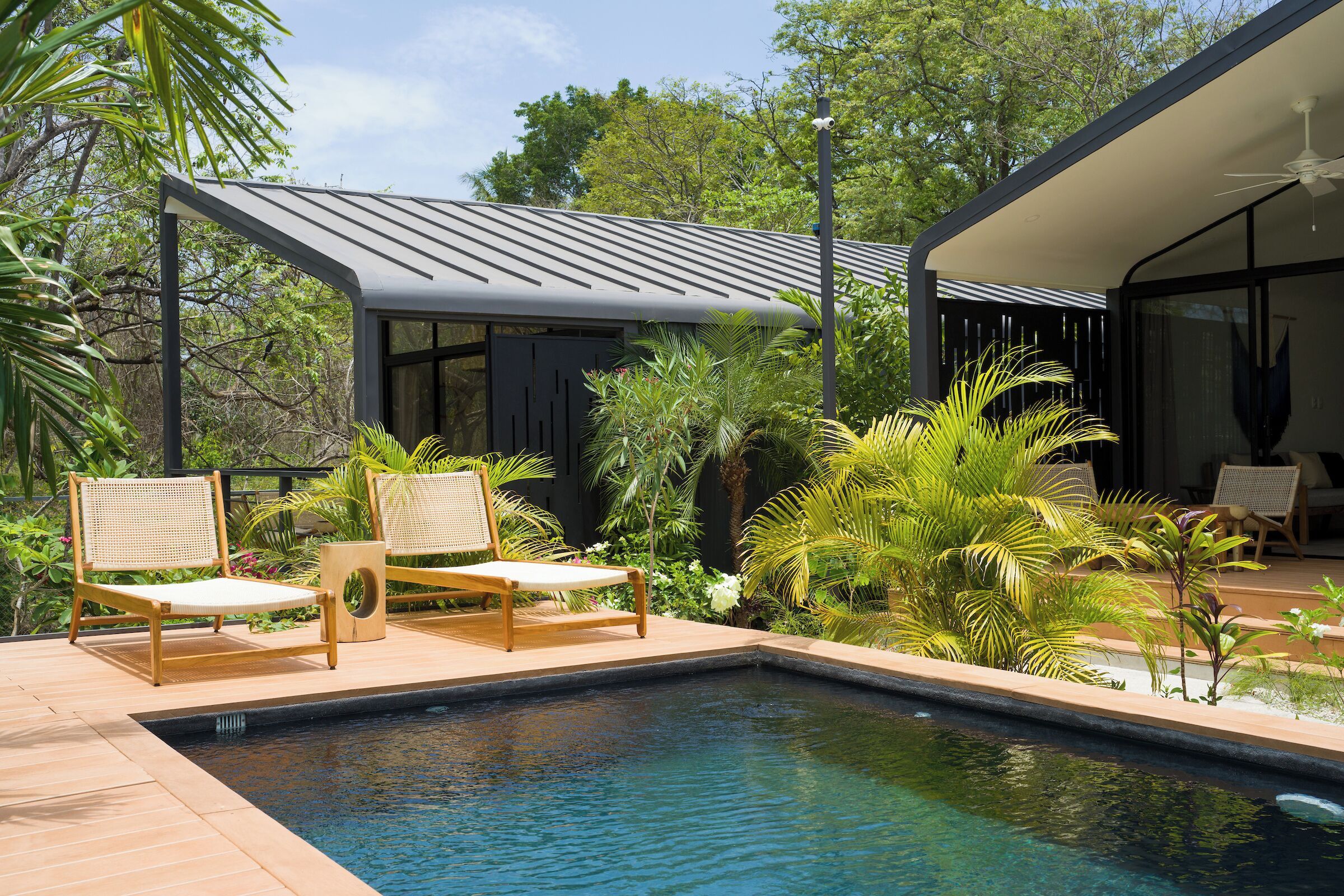 A modern outdoor pool area with two lounge chairs, a small building with a metal roof, and lush greenery.