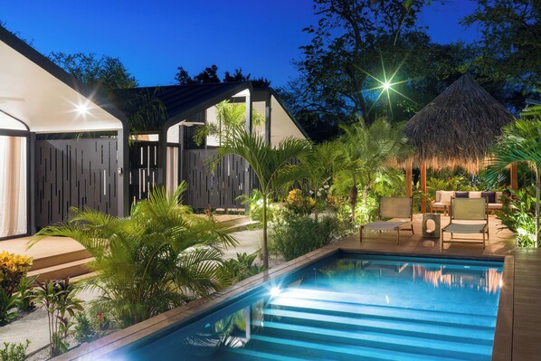 Un espace piscine extérieur moderne, doté d’une structure à toit de chaume, entouré d’une végétation luxuriante et agrémenté d’une terrasse en bois.