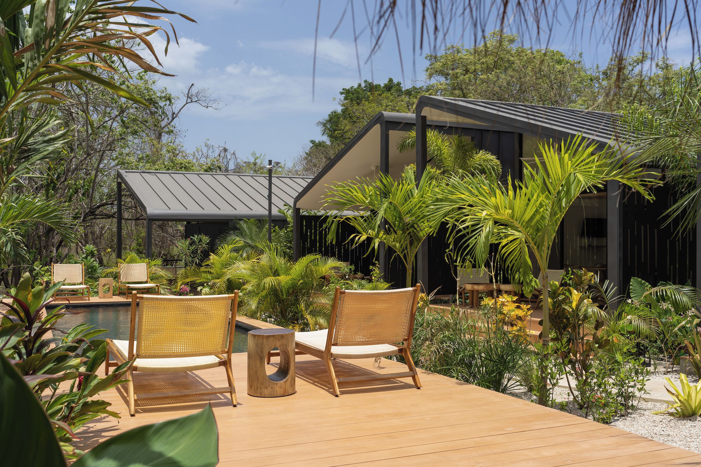 A wooden deck with wicker chairs, a small table, and a pool area surrounded by tropical plants.