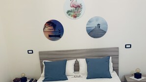 A neatly made bed with blue and white bedding, two bedside tables with lamps, and wall art depicting a flamingo, a sunset, and a pier. - All the Comforts of a Hotel With the Spaces of a Home (Castellammare di Stabia)