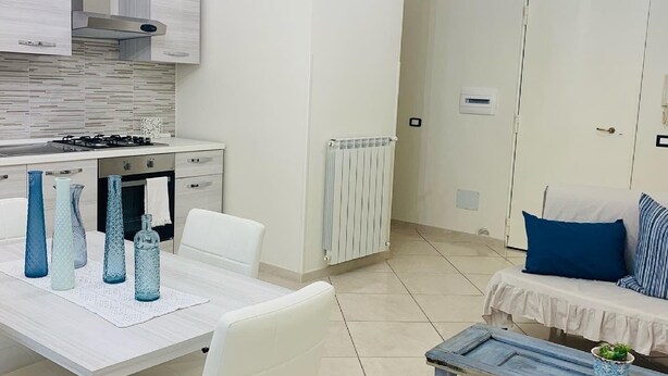 A modern kitchen with white cabinetry, a gas stove, and a white dining table with chairs. There is a white sofa with blue cushions and a small table with a plant. - All the Comforts of a Hotel With the Spaces of a Home (Castellammare di Stabia)