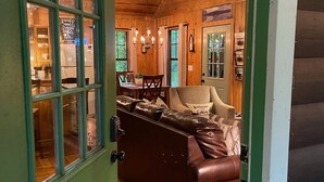 Een gezellig interieur van een chalet met een houten plafond, een eethoek met een kroonluchter, een leren bank en een groene deur.