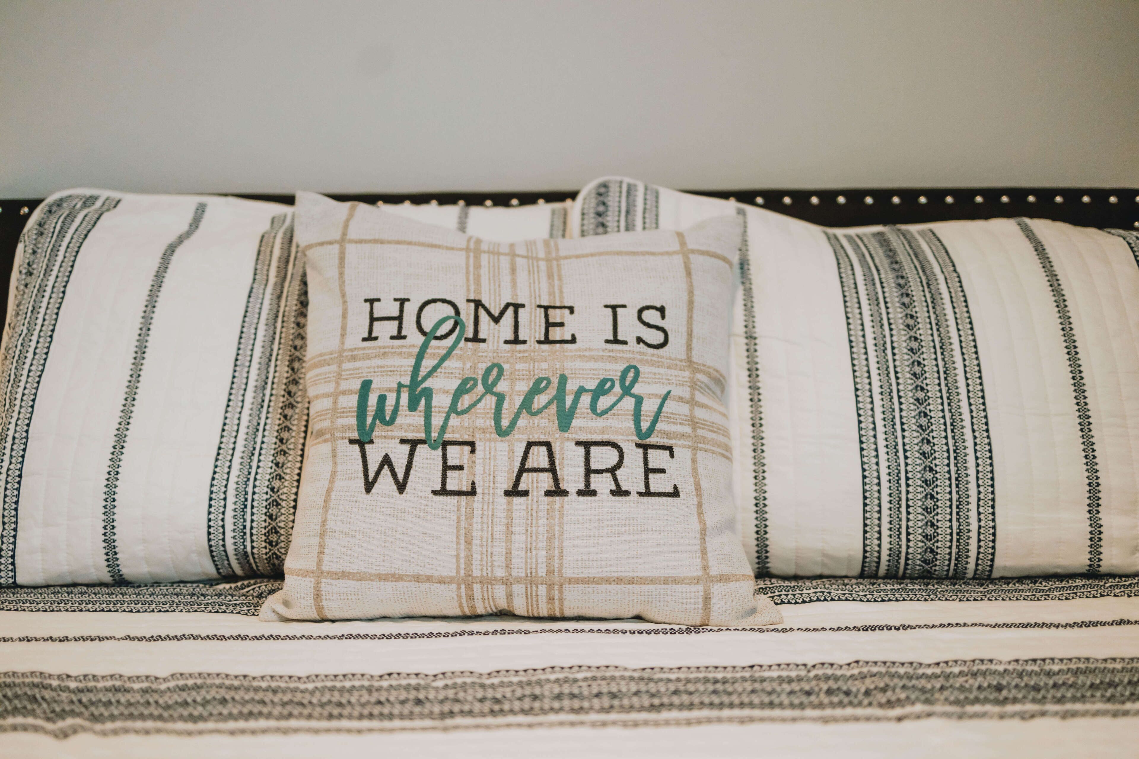 Un cuscino con la scritta "HOME IS WHEREVER WE ARE" su un letto con biancheria a righe.