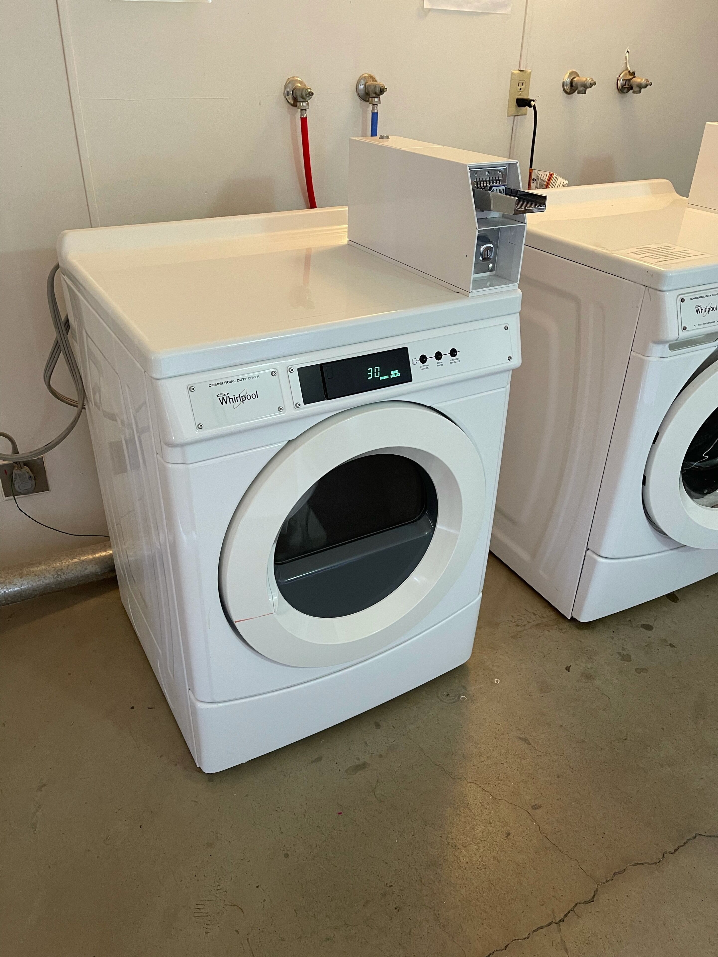 Dos lavadoras Whirlpool de carga frontal blancas en un lavadero con piso de concreto y un enchufe eléctrico instalado en la pared.