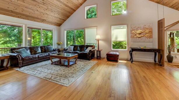 A spacious living room with a high wooden ceiling, large windows, a leather sofa, a coffee table, and a wooden floor. - Spacious Cabin with Hot Tub, Sauna, AC, Loft, King bed, Free Parking (Seattle)