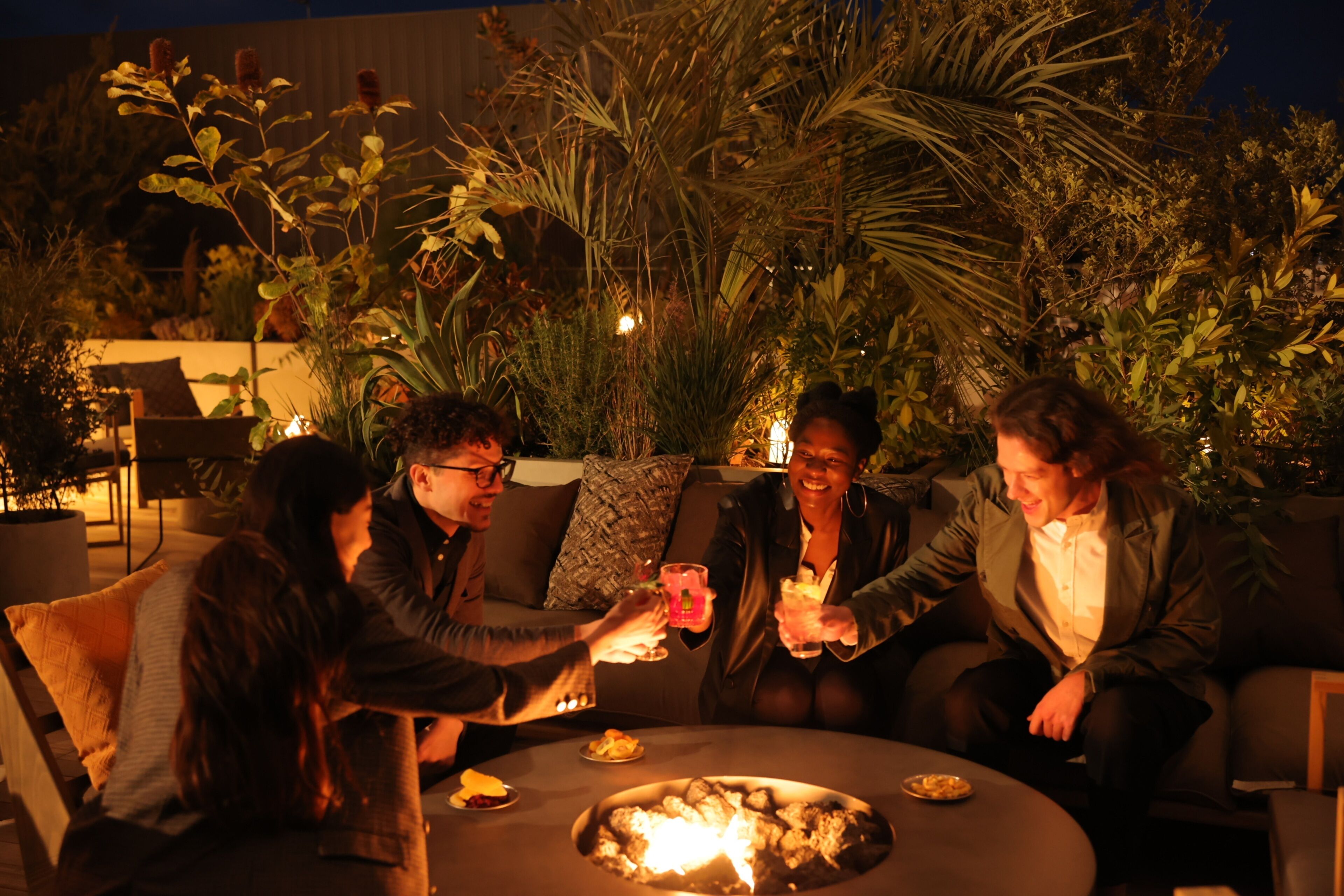 Un grupo de personas sentadas alrededor de una mesa redonda con una chimenea, rodeado de plantas y mobiliario de exterior.