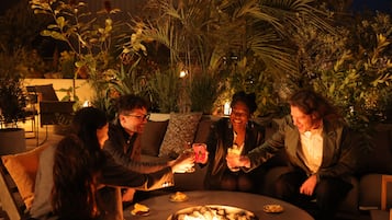 Un grupo de personas sentadas alrededor de una mesa redonda con una chimenea, rodeado de plantas y mobiliario de exterior.