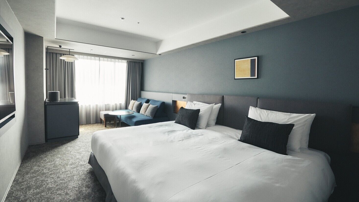 Quarto de hotel moderno com duas camas grandes, uma mesa de escritório, uma cadeira e uma janela ampla com cortinas.