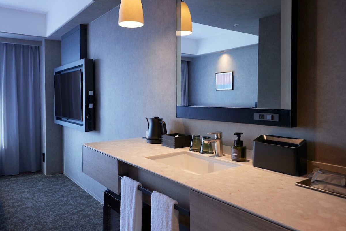 A modern hotel room with a large bed, a flat-screen TV, a large mirror, a sink with toiletries, and a coffee maker.