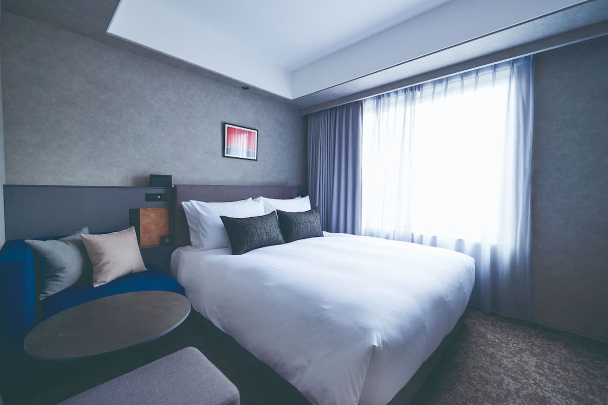 Une chambre d’hôtel moderne avec un grand lit, un canapé bleu, une petite table ronde et une grande fenêtre avec des rideaux.