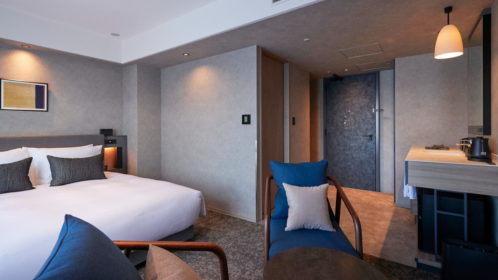 Quarto de hotel moderno com uma cama grande, área de estar com cadeiras, lareira e uma kitchenette pequena.