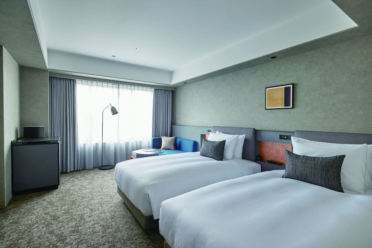 Une chambre d’hôtel avec deux lits, un bureau, une chaise, une lampe et une fenêtre avec des rideaux.