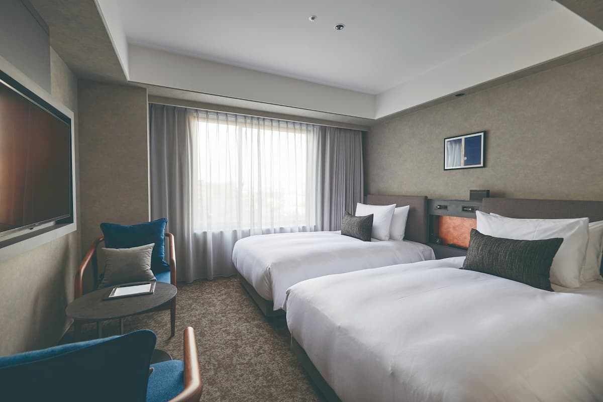 Une chambre d’hôtel avec deux lits, un bureau, une chaise et une grande fenêtre avec des rideaux.
