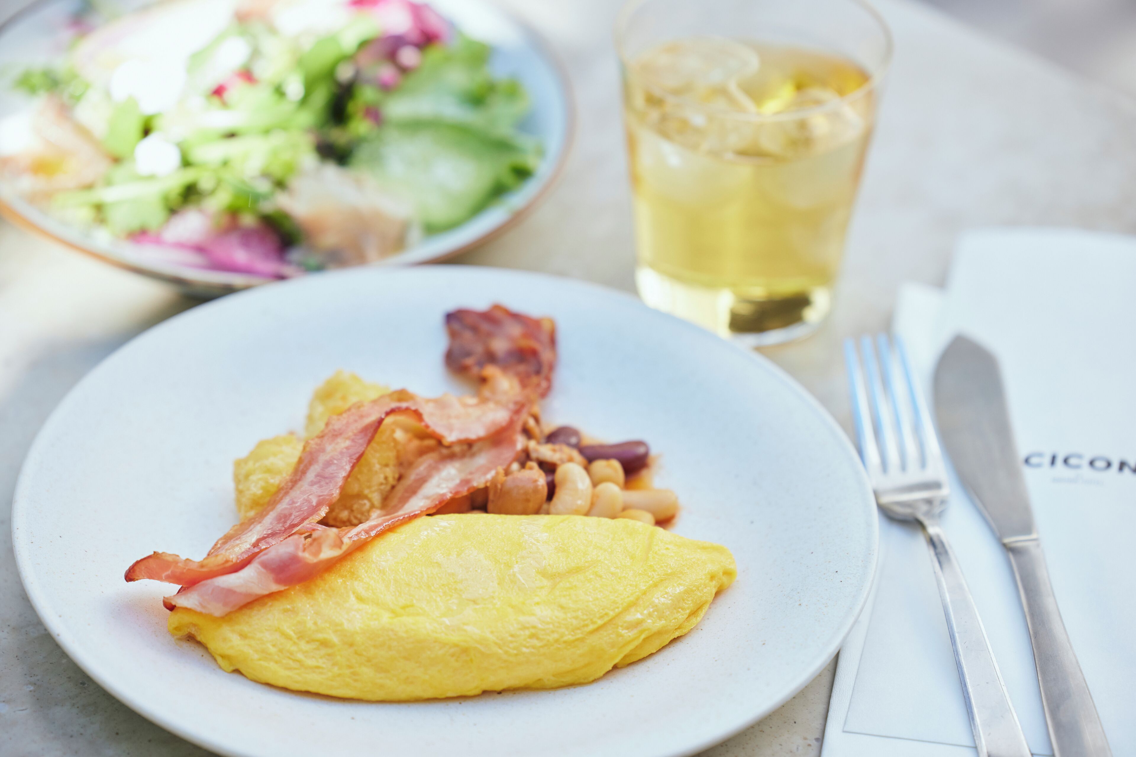 A plate with an omelet, bacon, and beans, a glass of iced tea, and a side salad on a table.