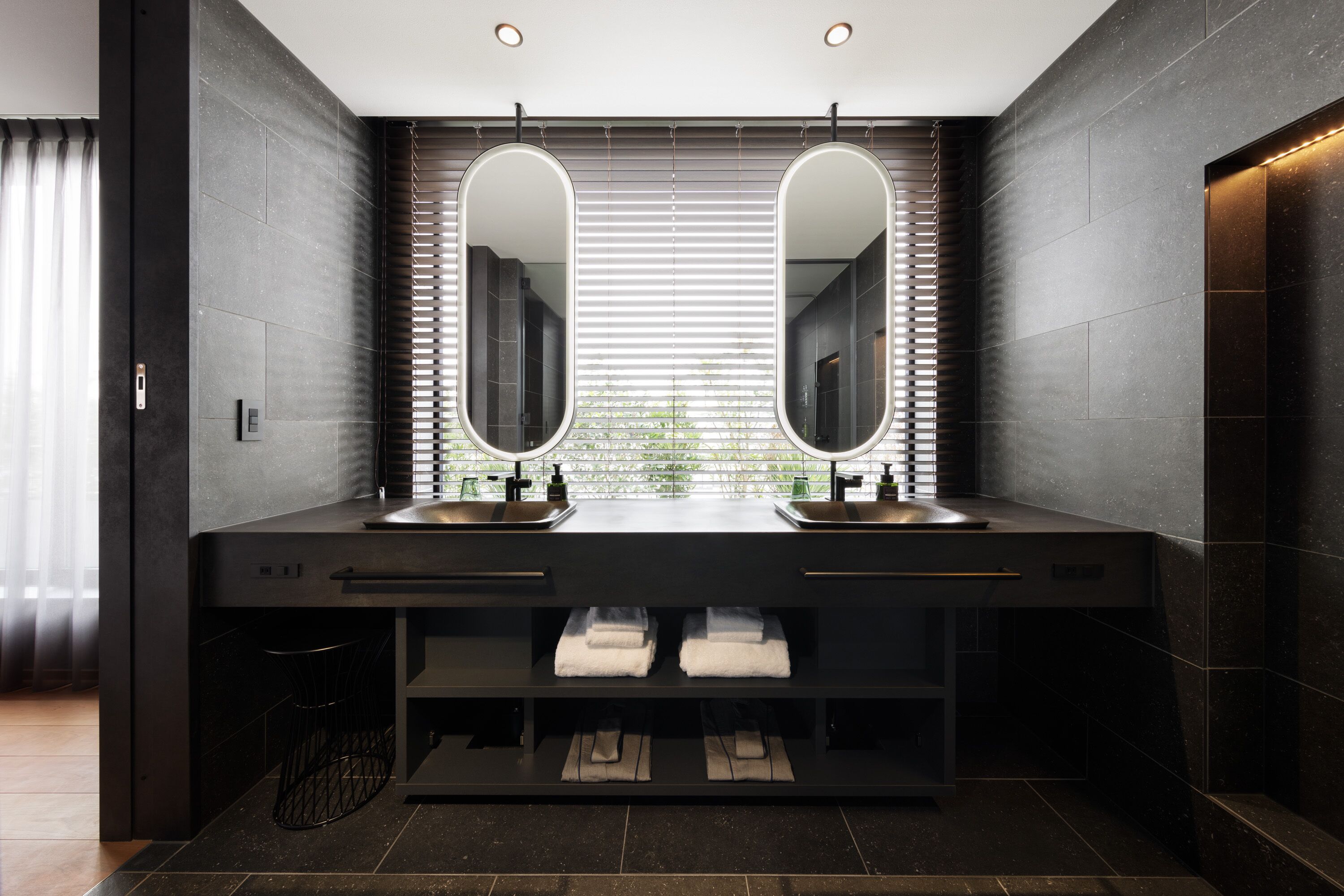 Une salle de bain moderne avec deux lavabos, des carreaux de couleur sombre et une fenêtre équipée de stores.