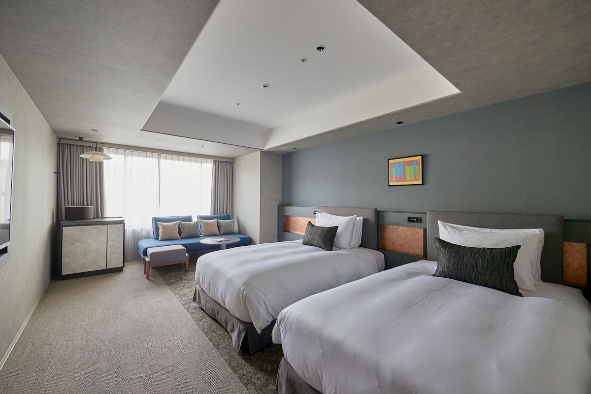 Une chambre d’hôtel moderne avec deux grands lits, un canapé, un bureau et une grande fenêtre avec des rideaux.