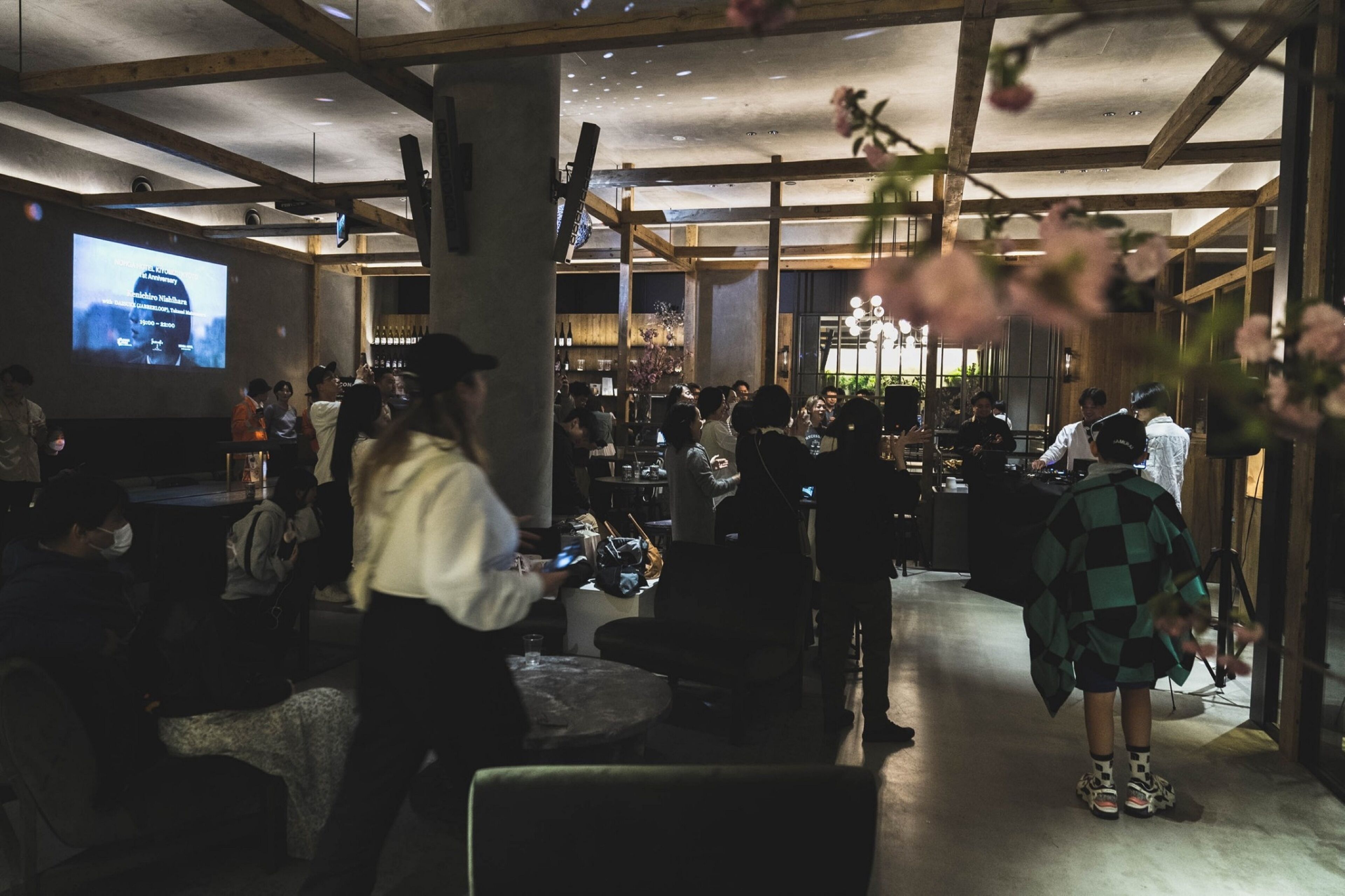 A crowded indoor event space with a large screen displaying a presentation, wooden beams on the ceiling, and a bar area in the background.