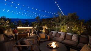 Un espacio al aire libre con fogata, sillones de descanso y luces colgantes, con montañas al fondo.