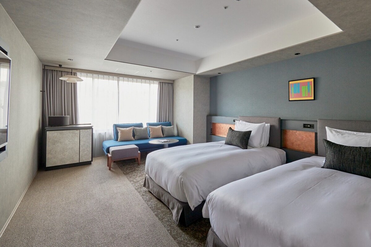 A hotel room with two beds, a sofa, a small table, and a view of the outdoors.