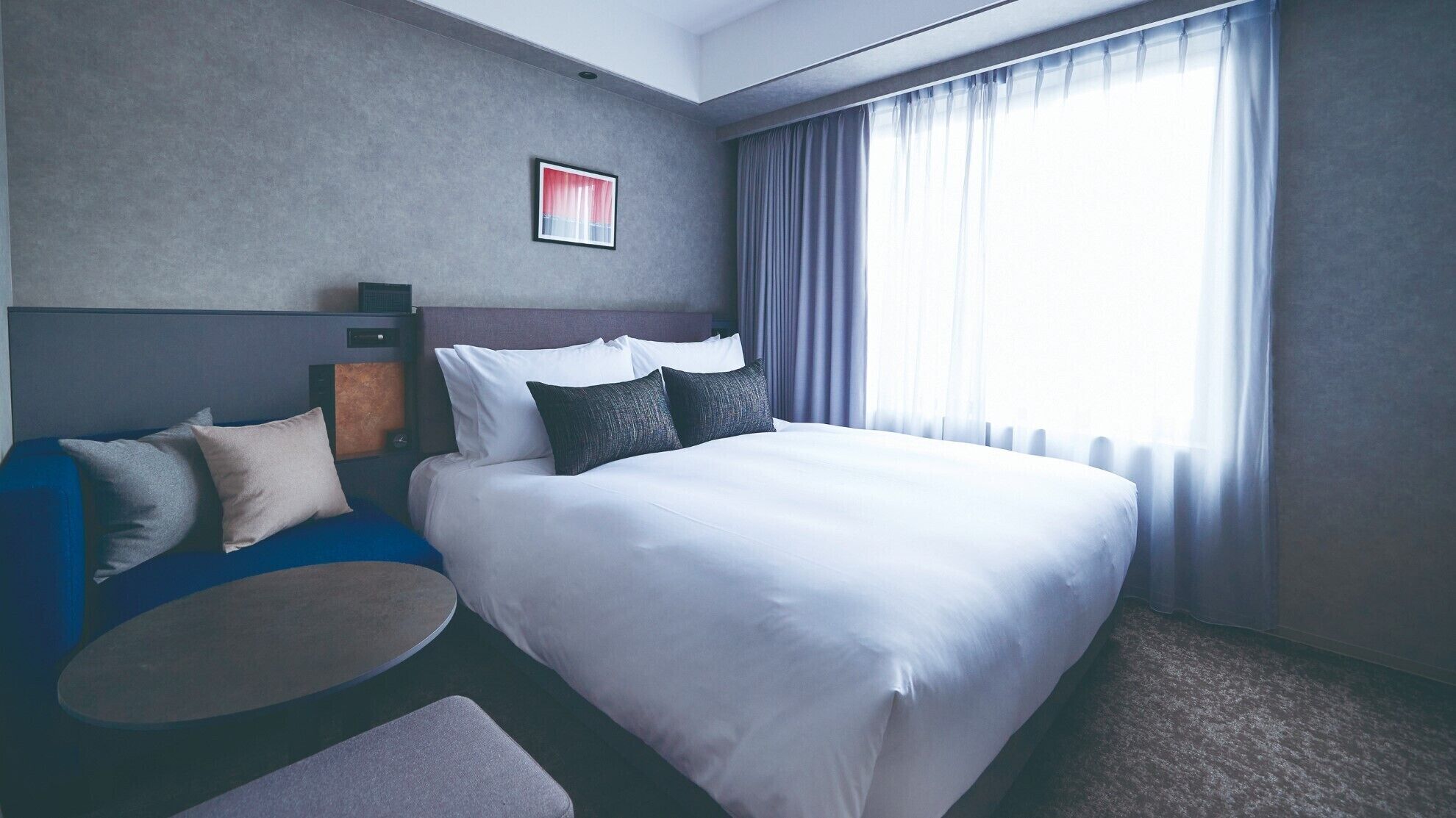 Quarto de hotel moderno com uma cama grande, área de estar com uma mesa redonda e uma janela ampla com cortinas.
